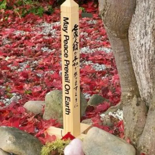 A wooden peace pole reading 'May Peace Prevail On Earth' set among stones and autumn leaves